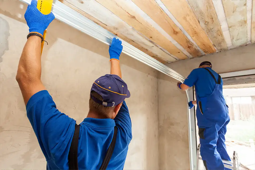 Zwei Handwerker in blauer Arbeitskleidung montieren gemeinsam eine Metallschiene an der Decke einer Garage. Einer hält die Schiene mit erhobenen Armen fest, während der andere auf einer Leiter steht und sie am Garagentorrahmen befestigt.